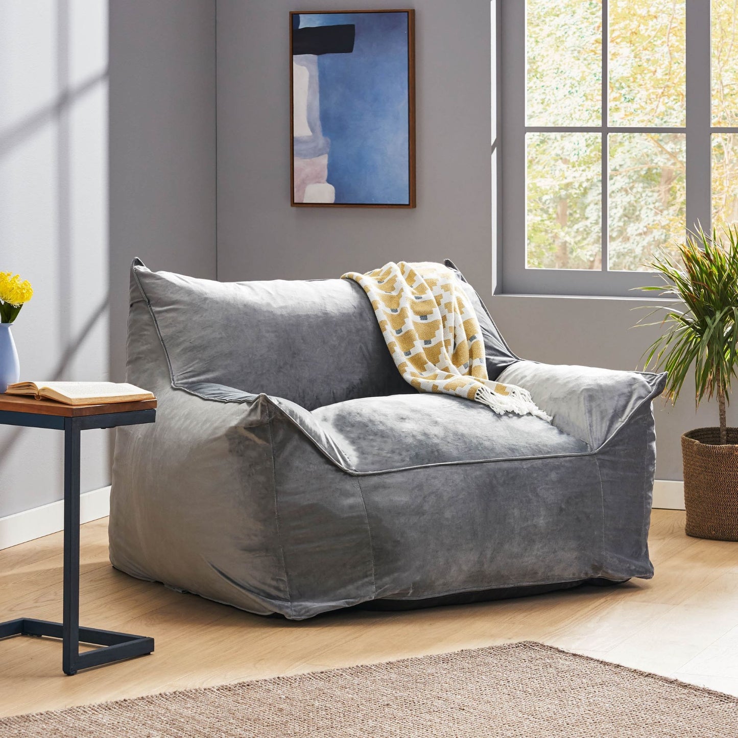 A cozy living room features the Allea Bean Bag Chair With Armrests in gray, draped with a yellow and white blanket, alongside a houseplant, small side table, rug, and a window with natural light.
