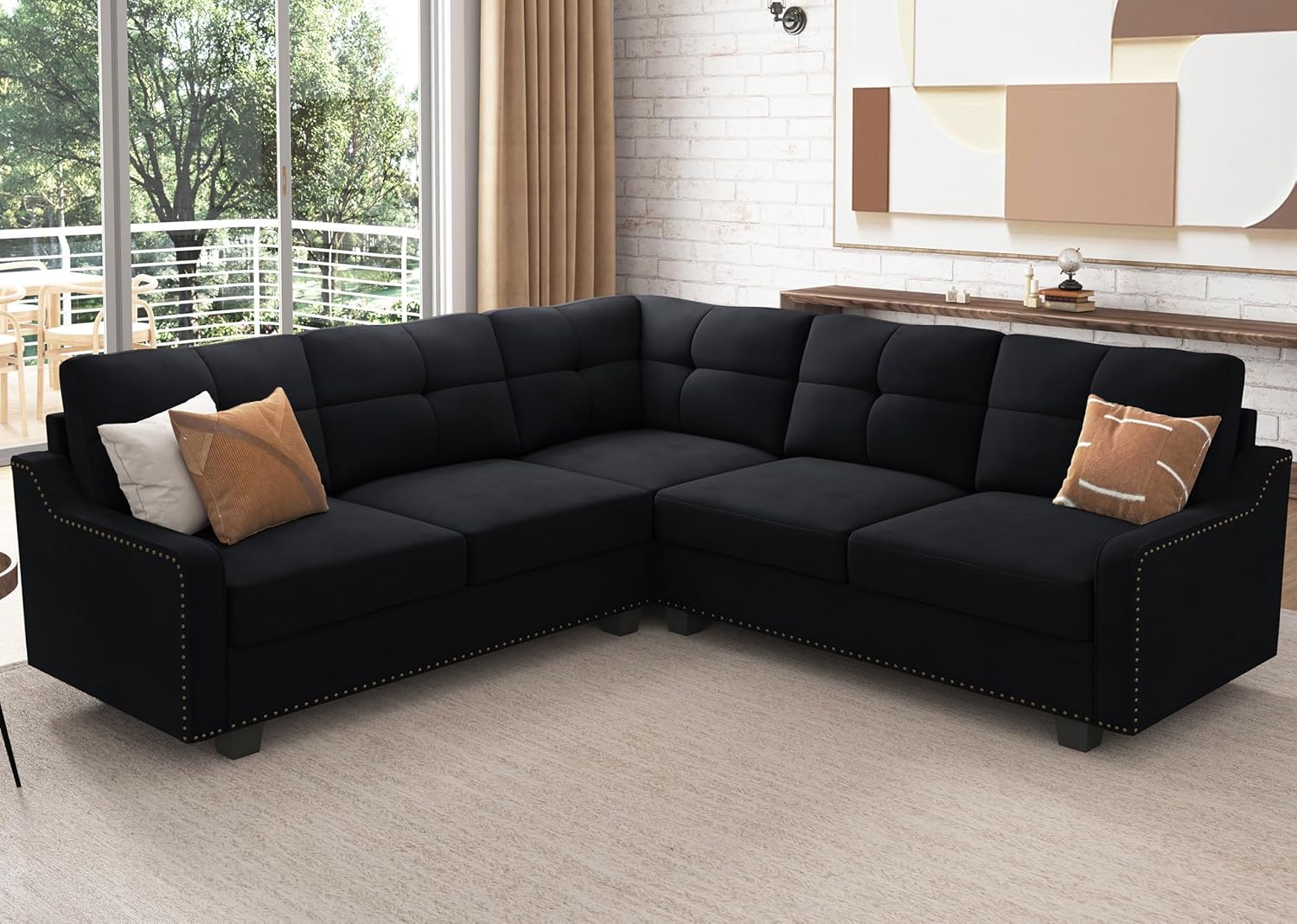 The stylish living room showcases the HONBAY Convertible Sectional Reversible Apartment, a black L-shaped sofa accented with button-tufted cushions and studded embellishments. It's elegantly decorated with several brown and beige throw pillows, while large windows provide a scenic view of trees and a balcony.