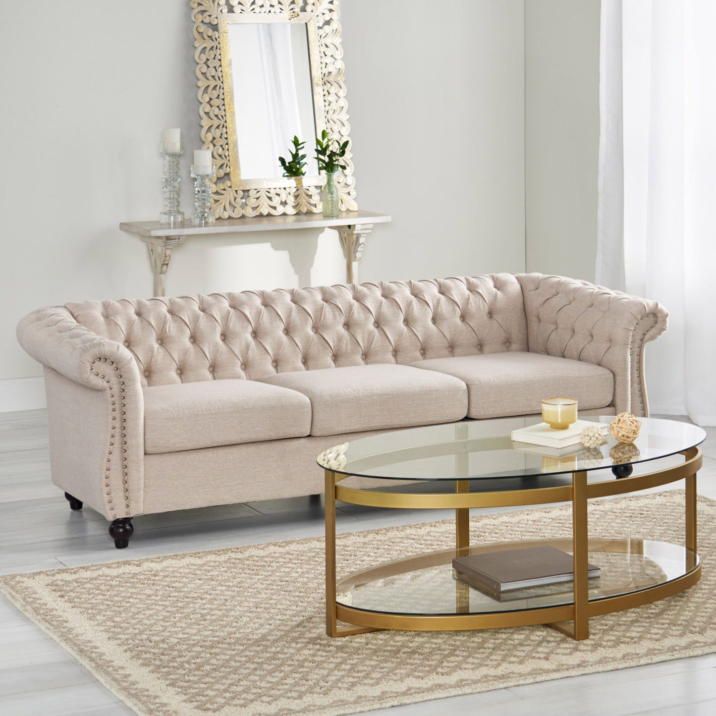 A Chesterfield Style 3 Seater Sofa with deep button tufting sits in a bright living room, paired with a gold-framed glass coffee table, decorative rug, wall mirror, candles, and potted plants on a shelf.
