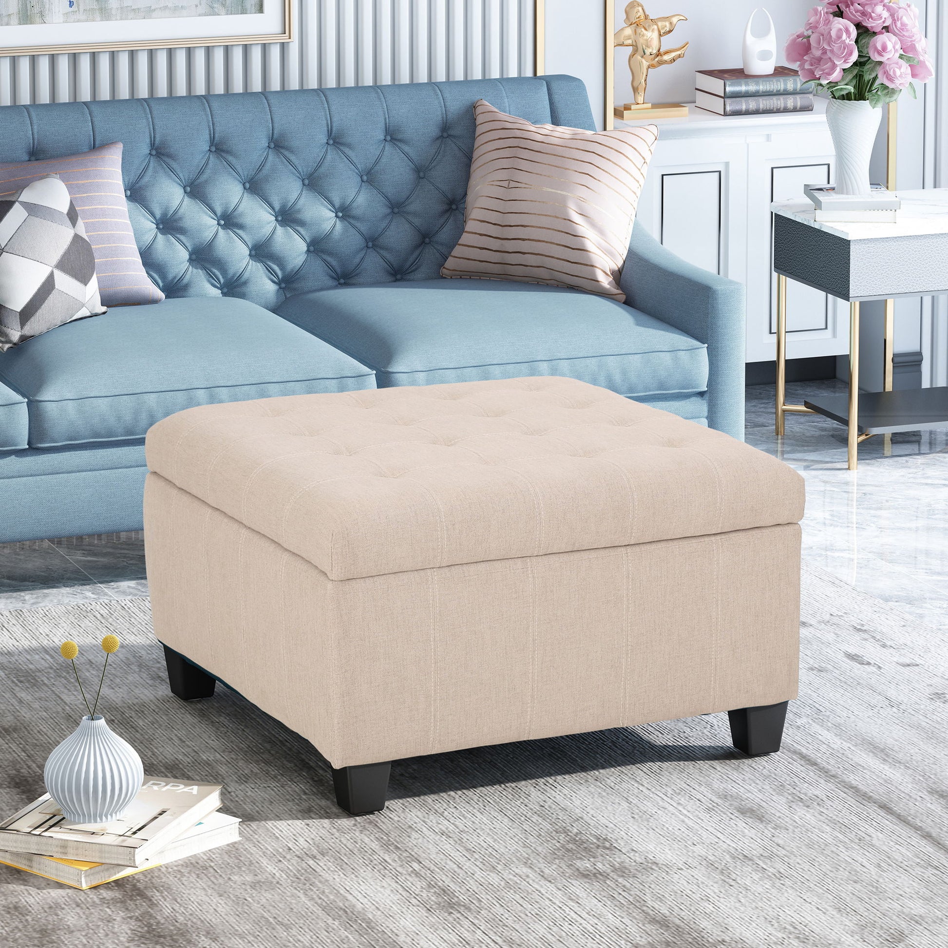 The Isabella Contemporary Ottoman With Storage, upholstered in beige with tufted detailing, sits before a blue tufted sofa adorned with decorative pillows in a modern, stylish living room.