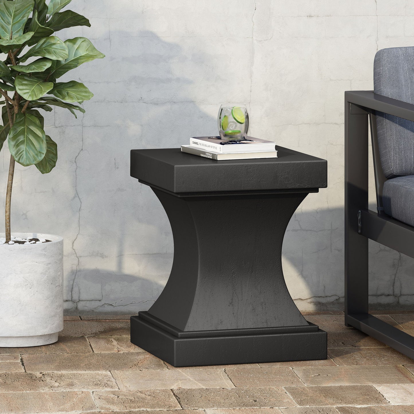 The Athena - Modern Side Table With Pedestal Design sits on a brick patio, holding books and a glass of lime water. A potted plant is to the left, and a cushioned outdoor chair appears partially on the right.