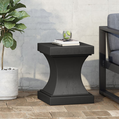 The Athena - Modern Side Table With Pedestal Design sits on a brick patio, holding books and a glass of lime water. A potted plant is to the left, and a cushioned outdoor chair appears partially on the right.
