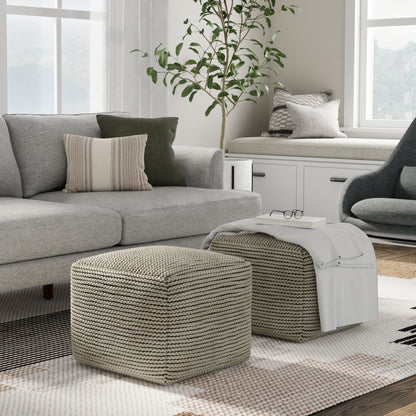 A modern living room with a gray sofa, patterned cushions, two Eastmont Outdoor/Indoor Poufs in cream, a cozy window seat, a plant, and a gray armchair. A white blanket and glasses rest on one pouf as sunlight streams through large windows.