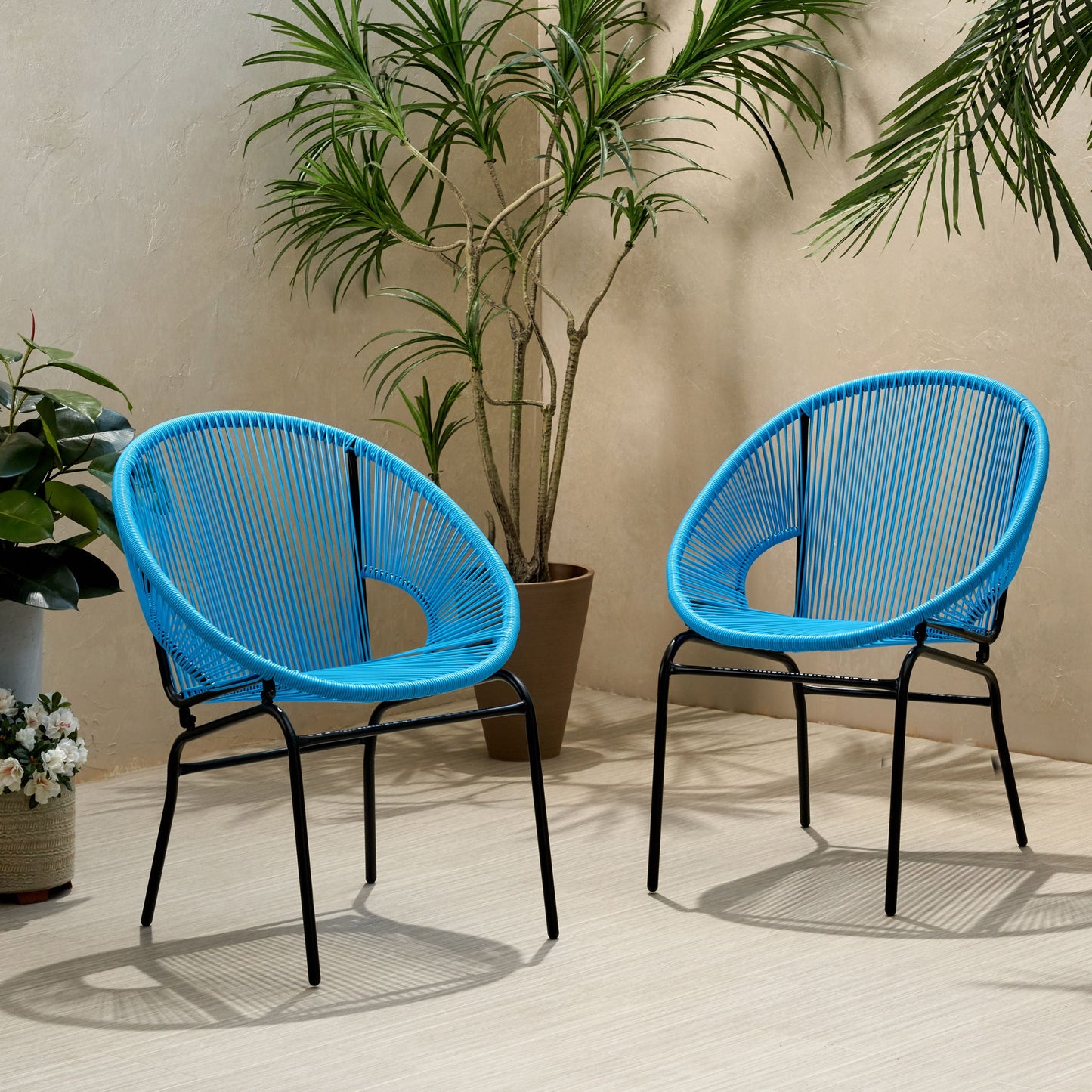 Two Nusa Modern Chair Outdoor Seating pieces with blue woven seats and black metal legs sit on a light floor, surrounded by green plants and a beige wall, creating a stylish, relaxed atmosphere.