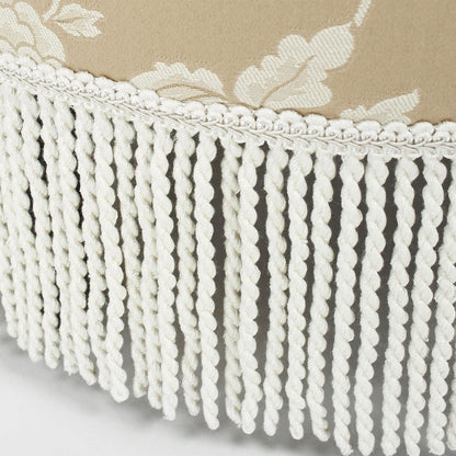 Close-up of the Yolanda Round Upholstered Accent Ottoman, featuring beige fabric with a white floral pattern and decorative white twisted fringe trim along the edge.
