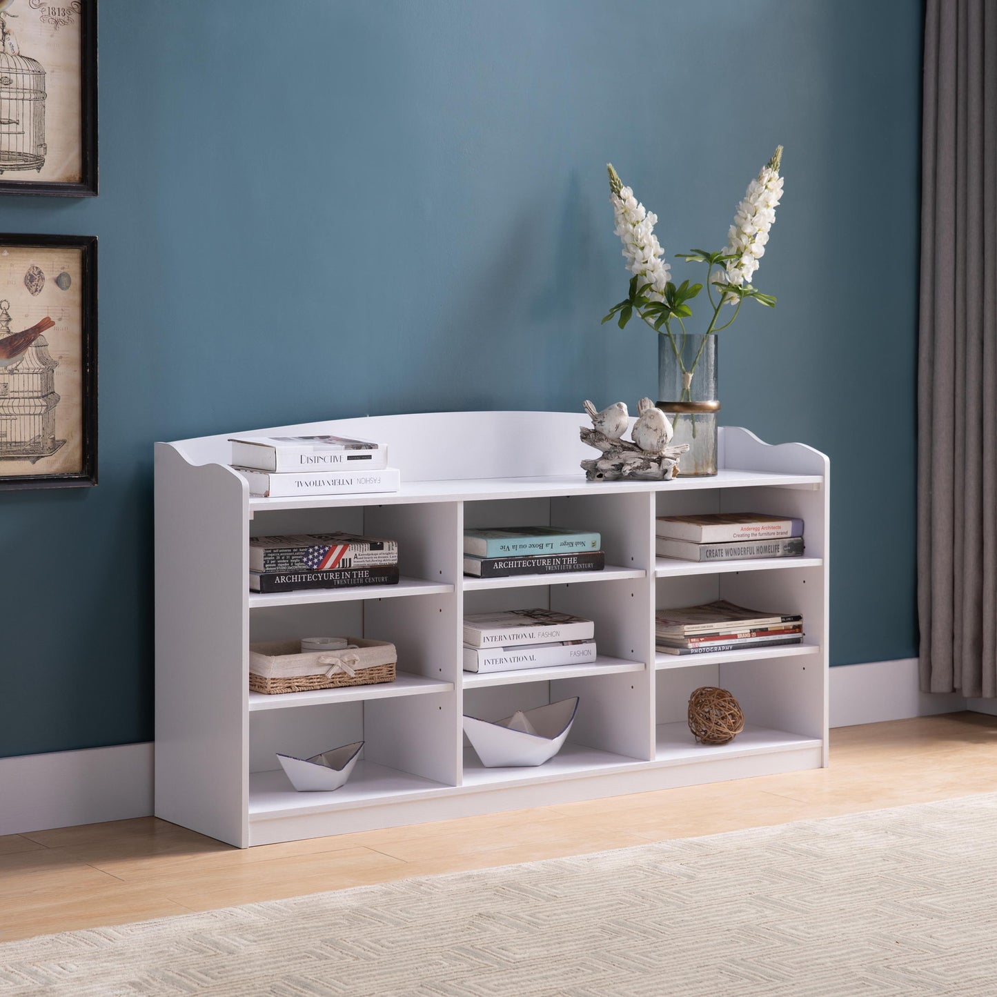 An Entryway Shoe Storage Bench with nine shelves holds shoes, baskets, and decor. A glass vase with white flowers and ceramic birds sits on top. The bench is set against a teal wall featuring artwork and a window curtain.