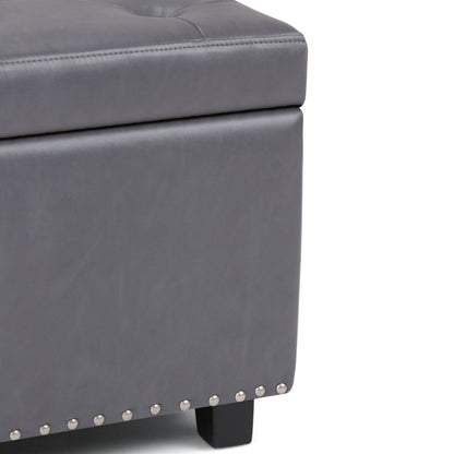 Close-up of the Hamilton Upholstered Storage Ottoman in gray, featuring silver nailhead trim, black wooden legs, and visible stitching on its tufted leather-like upholstery.