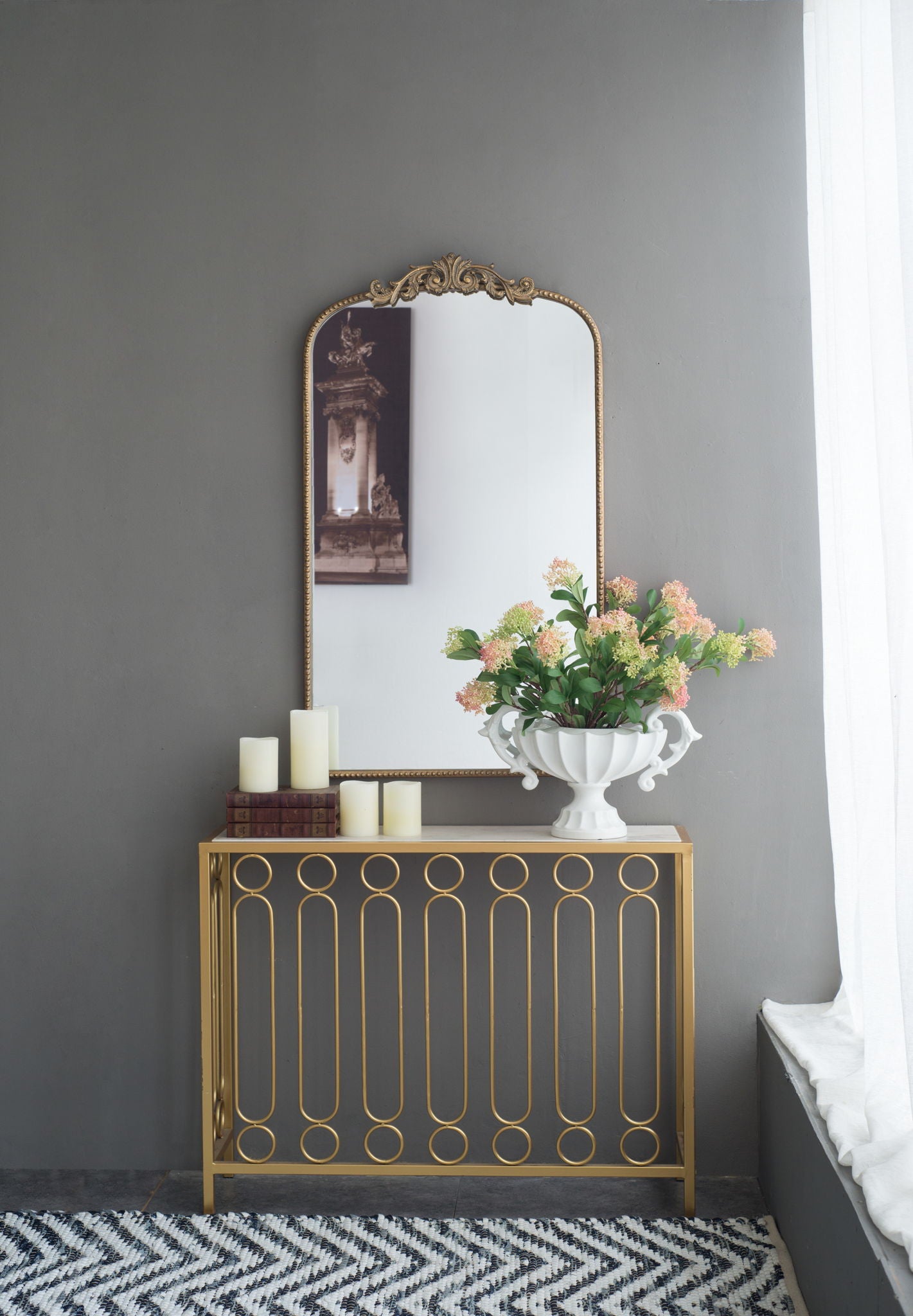 A gold console table with a white vase of flowers and pillar candles sits against a gray wall, topped by an Arch Mirror, Baroque Inspired Wall Decor for the living room. Sunlight filters through nearby white curtains.