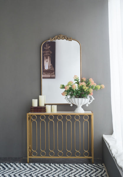 A gold console table with a white vase of flowers and pillar candles sits against a gray wall, topped by an Arch Mirror, Baroque Inspired Wall Decor for the living room. Sunlight filters through nearby white curtains.