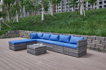The Patio Sofa With Cushions features a gray wicker L-shaped frame with bright blue cushions and a matching small table, set on a wooden deck near grass and trees.