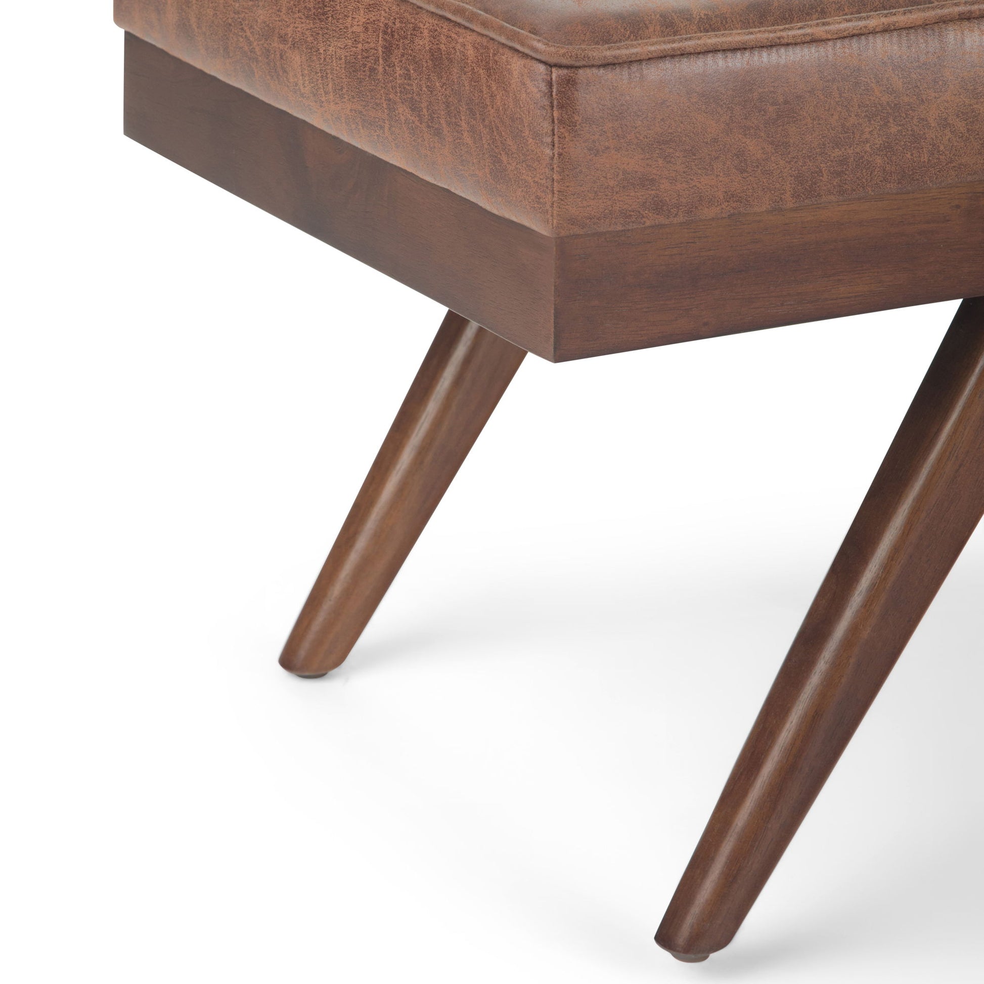 Close-up of the Pierce - Mid Century Upholstered Ottoman Bench featuring two angled wooden legs and a corner of its brown leather cushioned seat, set against a white background.