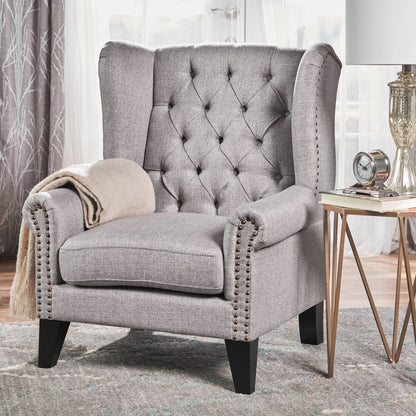 A Mid Century Modern Arm Reading Chair for the living room with tufted gray upholstery and nailhead trim sits on a rug with a beige throw blanket, near a side table holding a glass lamp, clock, and book. Curtains frame the window in the background.