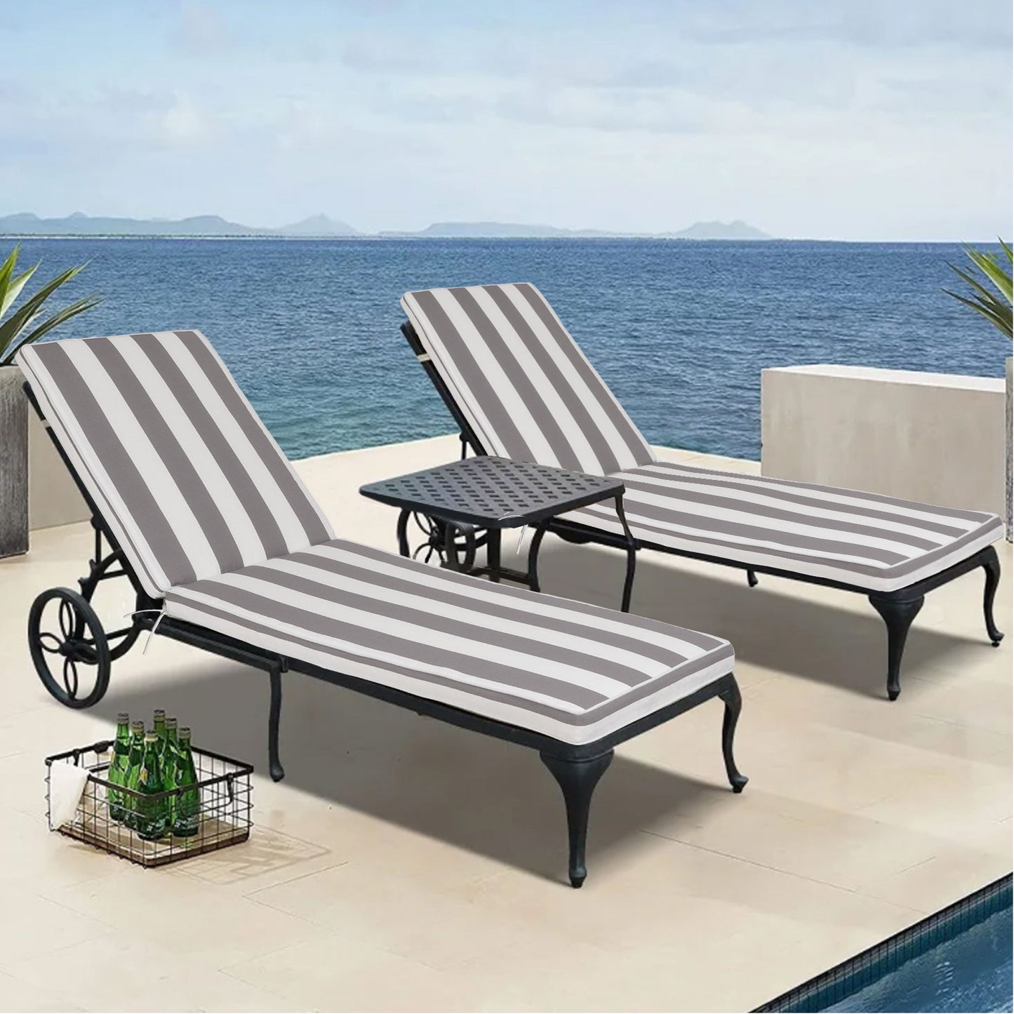 Two striped Outdoor Lounge Chair Cushion Replacement Patio Furniture Seat Cushions sit on lounge chairs by a small patio table, overlooking the ocean with distant islands visible and a wire basket of bottled drinks nearby.