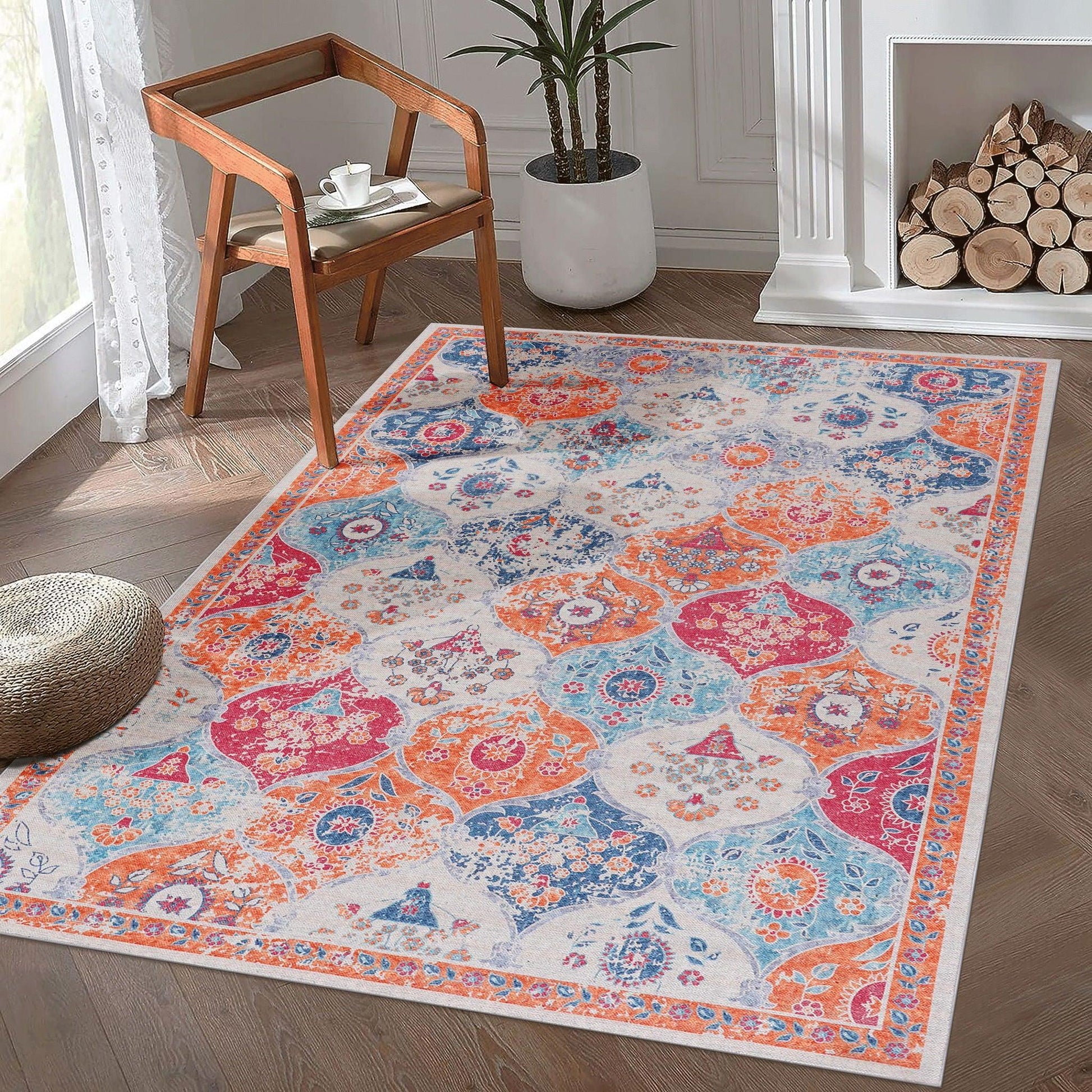 The Maval - Area Rug, featuring blue, orange, and red patterns on a light background, is displayed in a bright room with wood flooring, a wooden chair, potted plant, woven basket, and a fireplace with stacked logs.