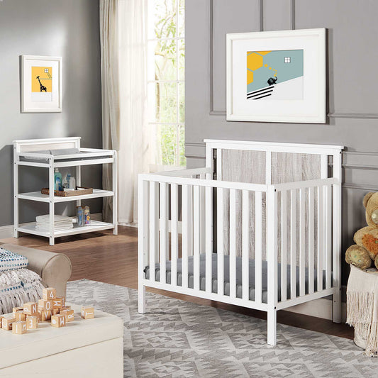 A modern nursery featuring the Connelly Mini Crib with Mattress Pad, a matching changing table, plush rugs, stuffed animals, wooden blocks, and framed artwork on gray walls by a large window with natural light.
