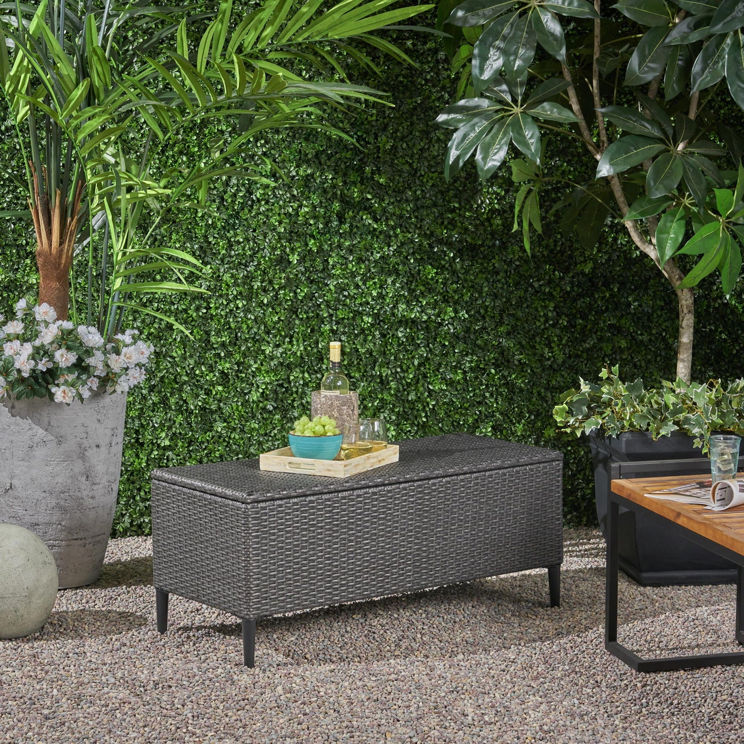 The Rupert - Storage Unit Resin Wicker Outdoor sits on gravel, topped with a tray holding a bottle, glasses, and a bowl. Lush green plants and trees surround the area, giving it a garden-like ambiance.