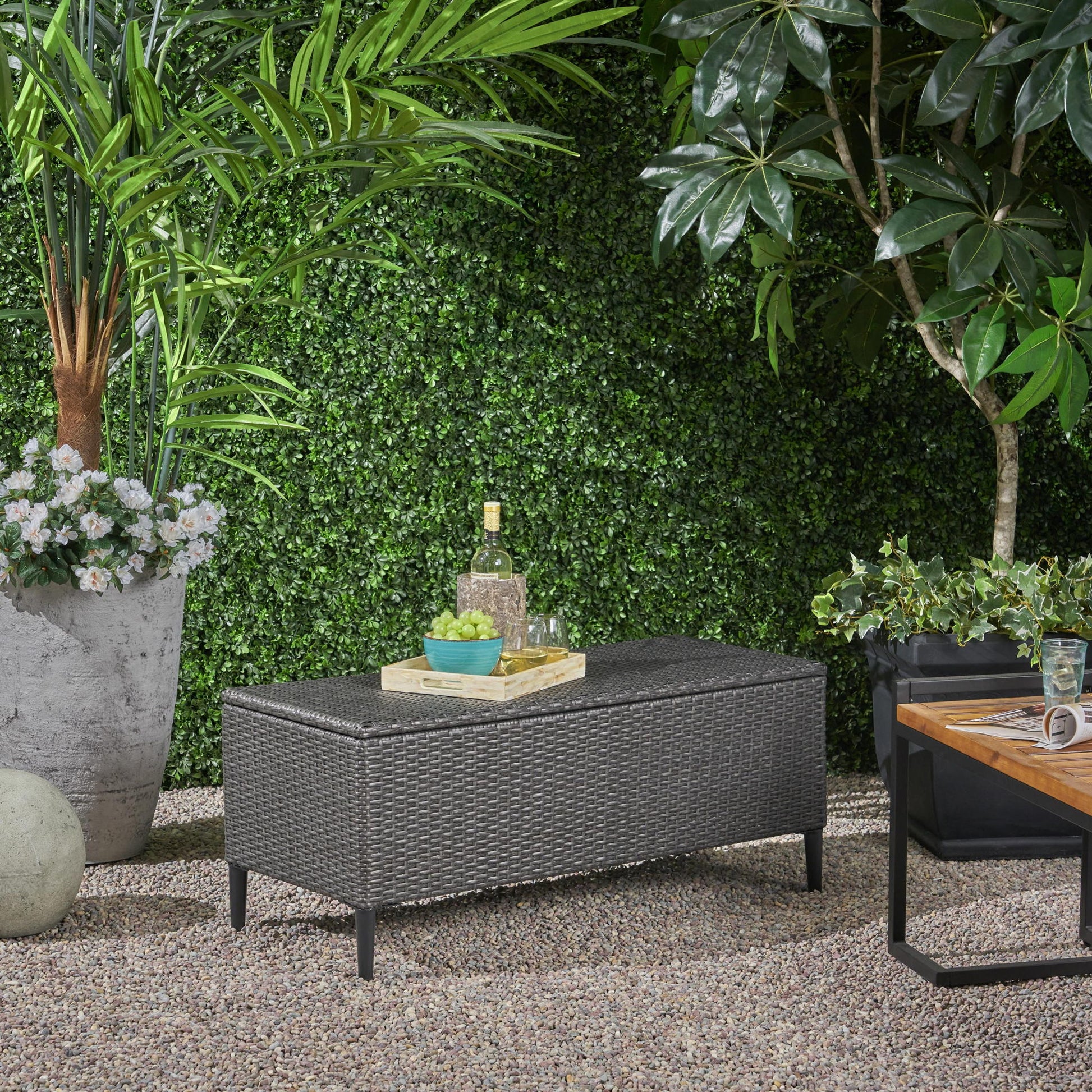 The Rupert - Storage Unit Resin Wicker Outdoor sits on gravel, topped with a tray holding a bottle, glasses, and a bowl. Lush green plants and trees surround the area, giving it a garden-like ambiance.