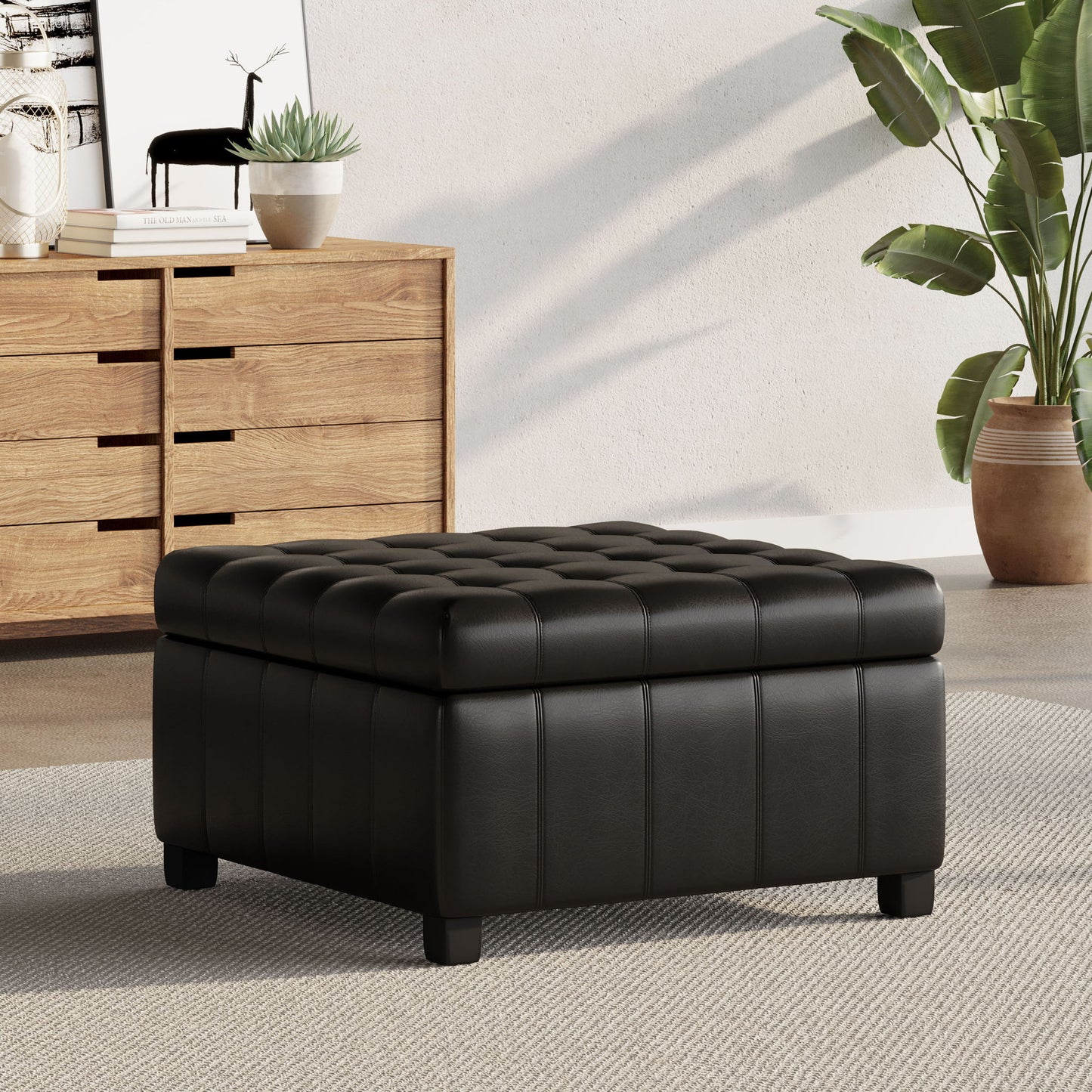 A Storage Ottoman For Living Room in black tufted leather sits on a light rug, complemented by a wooden dresser, potted plants, and framed art in this modern living space.