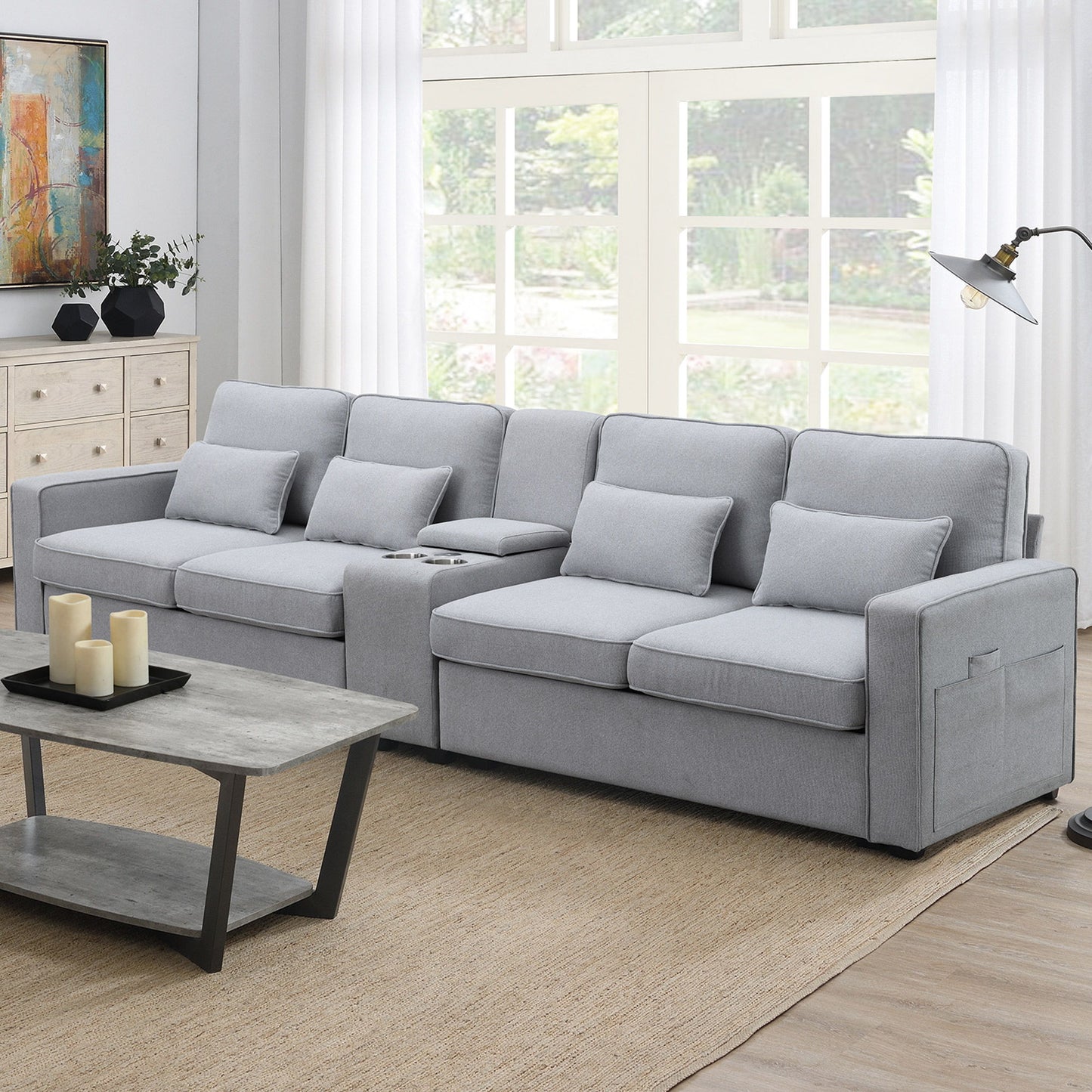 A modern living room with a large light gray Upholstered Sofa with console, 2 cupholders, 2 USB ports (wired/wireless charging), linen fabric and 4 pillows, paired with a wooden coffee table, beige rug, floor lamp, and big windows filling the space with natural light.