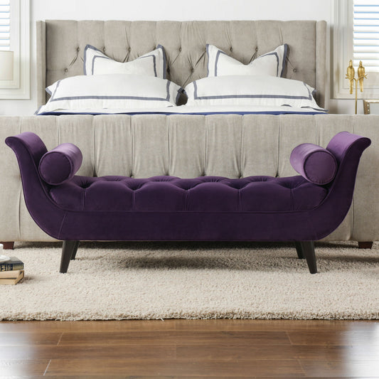 The Alma - Tufted Flared Arm Entryway Bench in deep purple velvet with curved arms and black legs sits at the foot of a beige upholstered bed with white pillows in a modern bedroom.