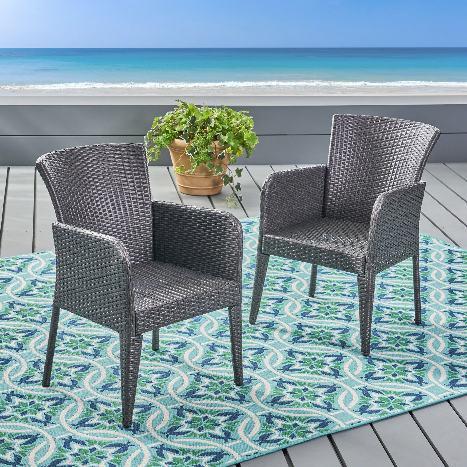 Two Anaya Outdoor Wicker Dining Armchairs (Set of 2) are arranged on a blue and green patterned rug on a deck overlooking the ocean, with a potted plant placed behind them.