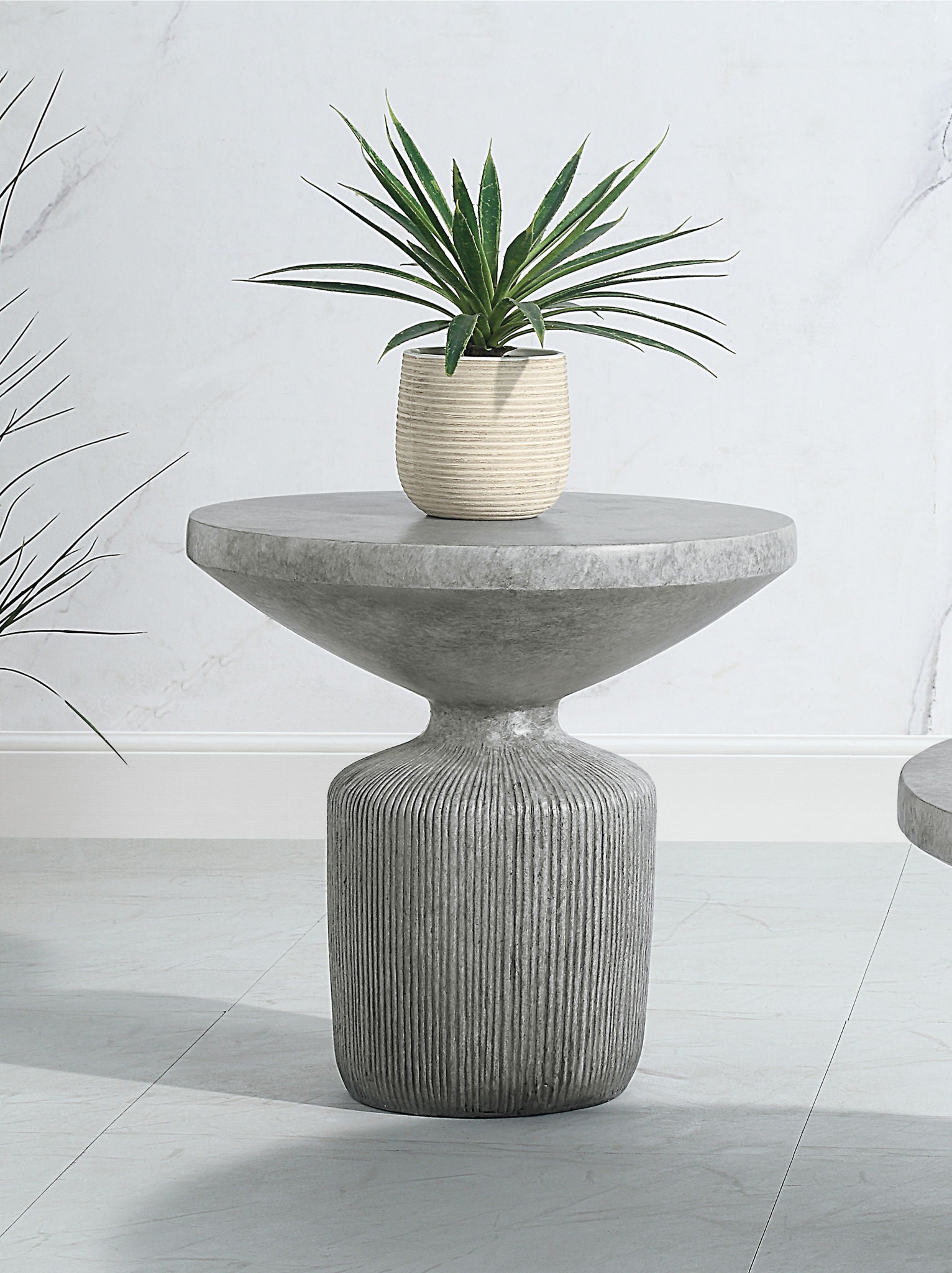 The Laddie - Weathered Table is a modern round concrete side table with a textured pedestal base, shown with a small potted green plant on top against a light-colored wall and floor.
