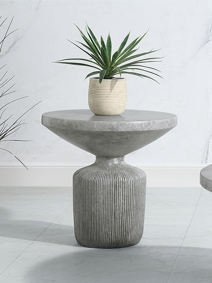 The Laddie - Weathered Table is a modern round concrete side table with a textured pedestal base, shown with a small potted green plant on top against a light-colored wall and floor.