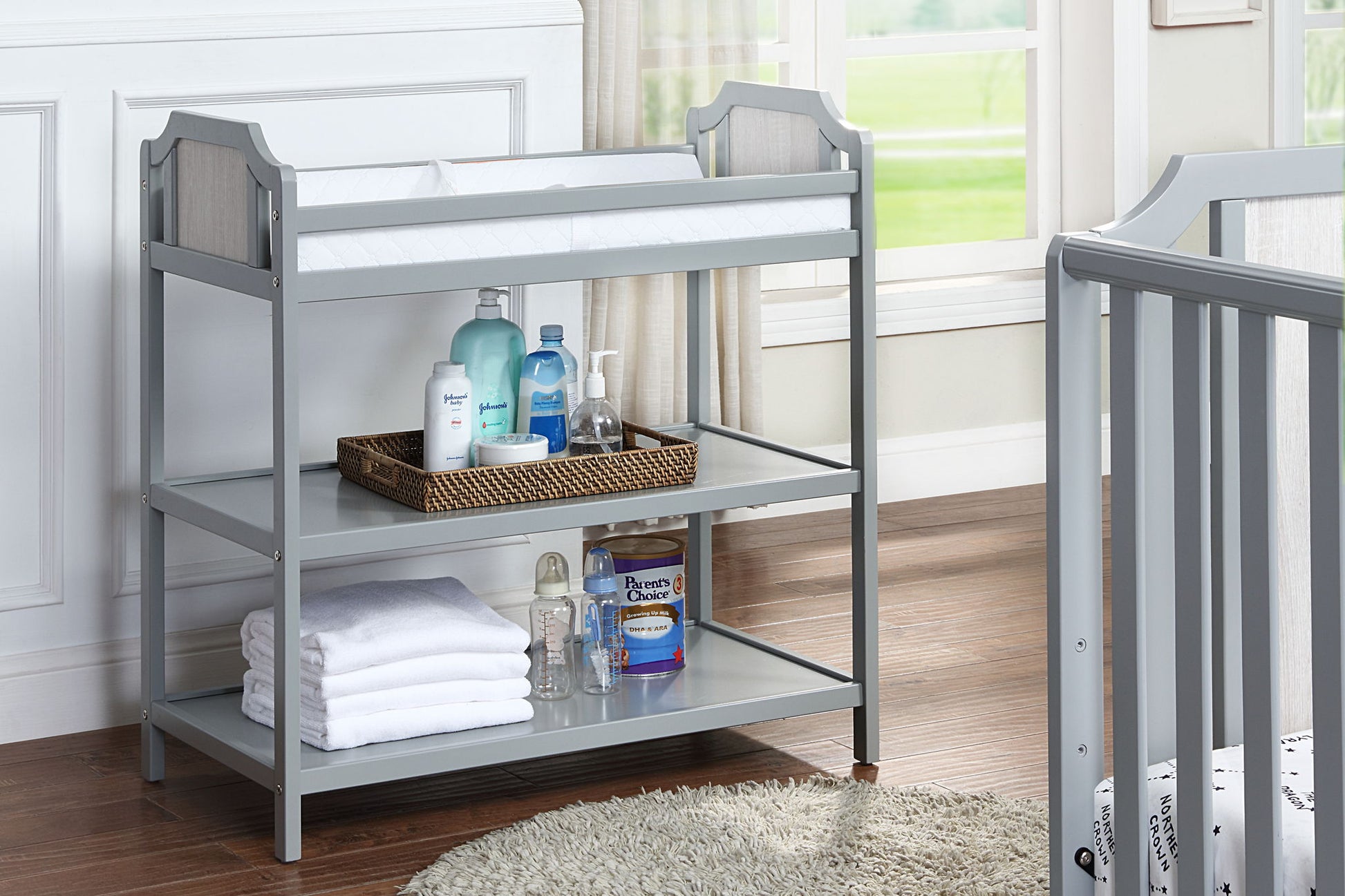 The Brees - Changing Table in gray, featuring a white changing pad, stacked towels, and baby care products on its shelves, stands in a bright nursery with wooden floors and a round rug.