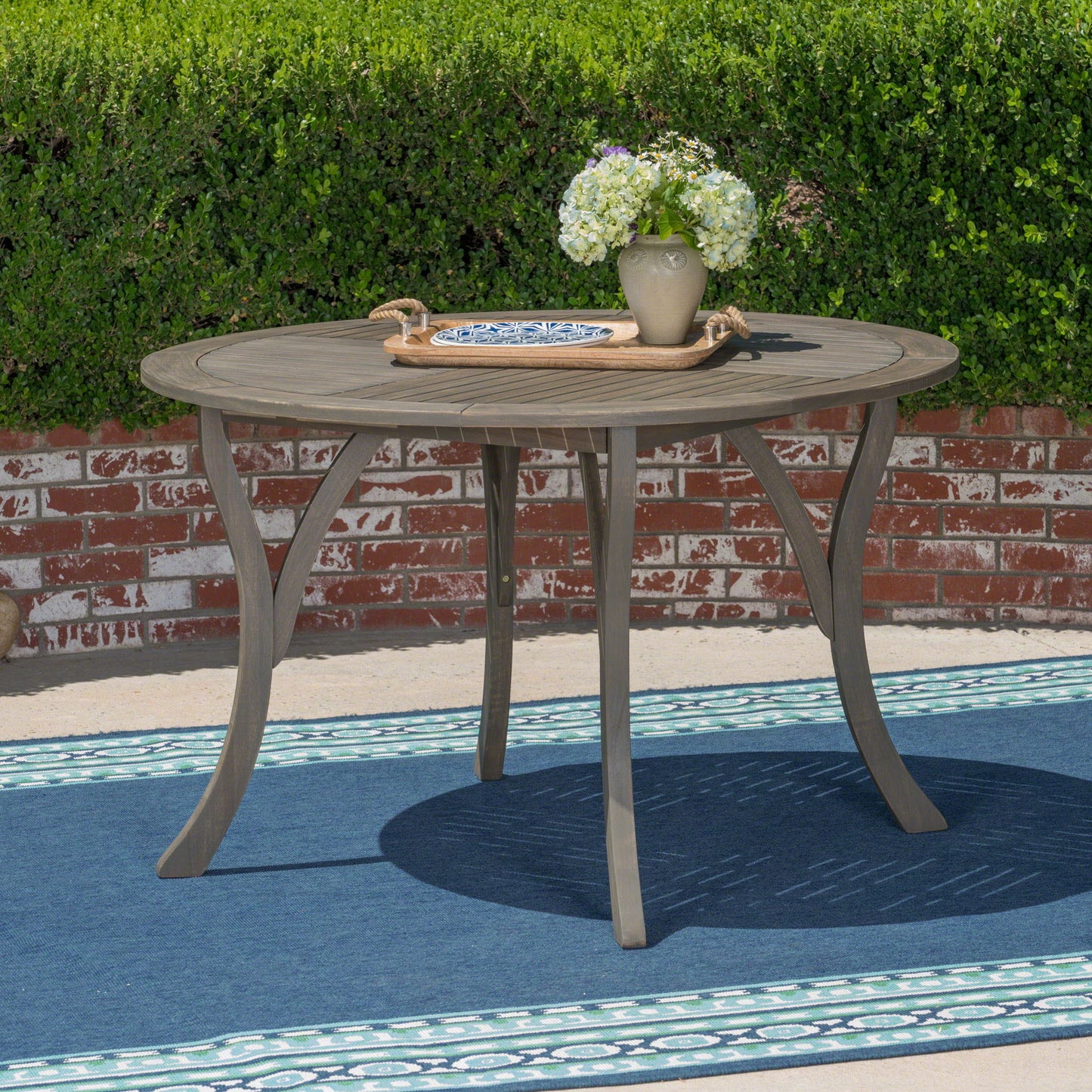 The Hermosa Outdoor Acacia Wood Table with a circular design and curved legs sits on a blue rug, topped with a tray holding a vase of flowers. A brick wall and green hedge form the background.