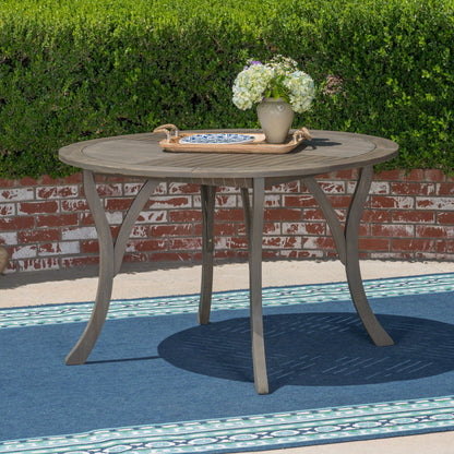 The Hermosa Outdoor Acacia Wood Table with a circular design and curved legs sits on a blue rug, topped with a tray holding a vase of flowers. A brick wall and green hedge form the background.