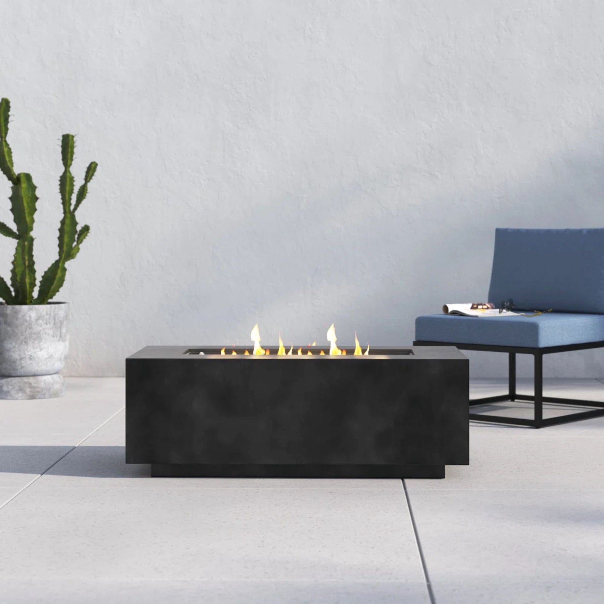 A modern patio features a Modern Concrete Design Propane Outdoor Fire Pit Table with visible flames at the center, a potted cactus on the left, and a blue cushioned chair on the right, all set against a light wall.