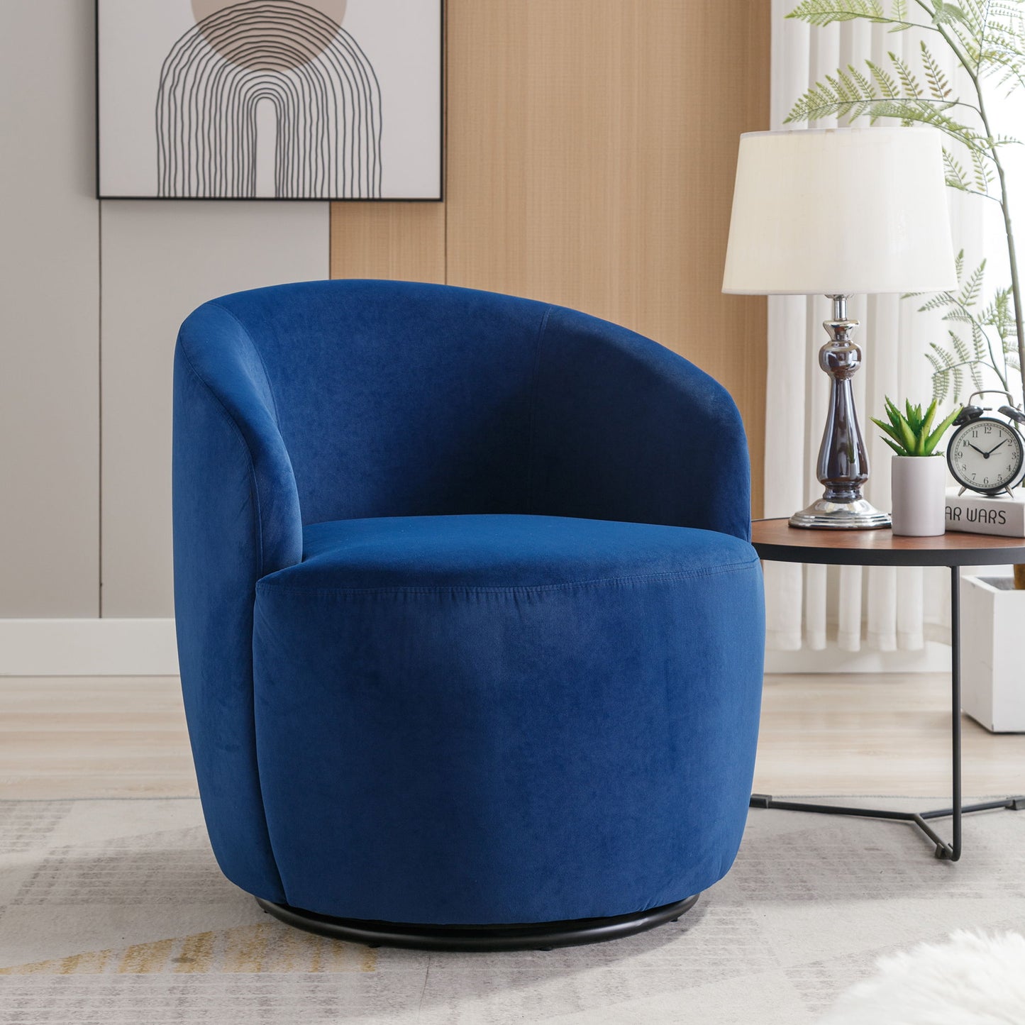 A modern living room showcases the Velvet Fabric Swivel Accent Armchair Barrel Chair With Powder Coating Metal Ring in blue, placed beside a side table with a lamp, clock, and books. A framed abstract art and potted plant complete the background.