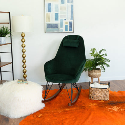 A modern living space with the Chloe Mid Century Modern Rocker chair in dark green, a fluffy white ottoman, gold floor lamp, potted plant, woven basket, and abstract wall art atop an orange rug on wood flooring.