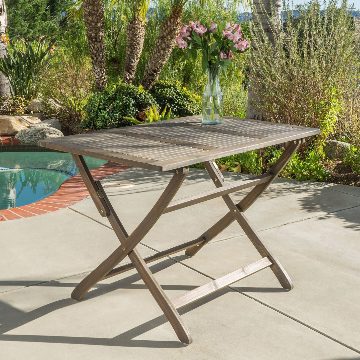 The Blissique Outdoor Foldable Wood Dining Table with a slat design stands on a sunny patio near a pool, topped with a glass vase filled with pink flowers and surrounded by greenery and palm trees.
