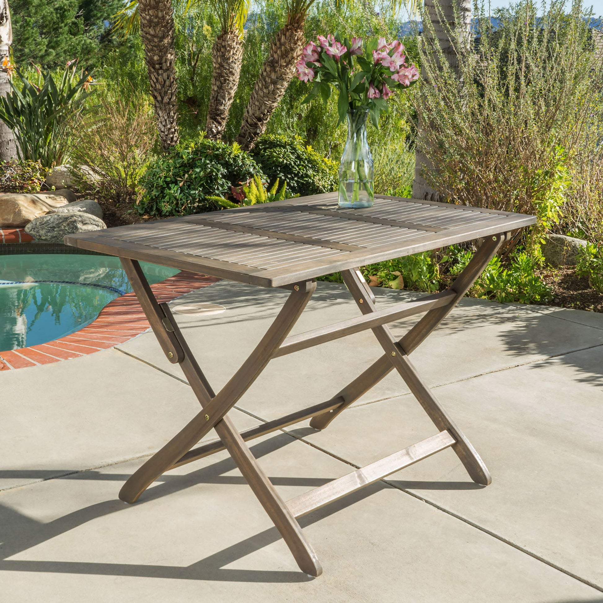 The Blissique Outdoor Foldable Wood Dining Table with a slat design stands on a sunny patio near a pool, topped with a glass vase filled with pink flowers and surrounded by greenery and palm trees.