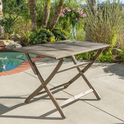 The Blissique Outdoor Foldable Wood Dining Table with a slat design stands on a sunny patio near a pool, topped with a glass vase filled with pink flowers and surrounded by greenery and palm trees.