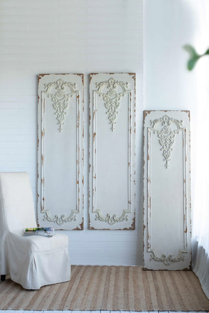 Three large wooden rectangle hanging panels with intricate designs lean against a white brick wall. A slipcovered chair on a woven rug holds a stack of books, while natural light fills the space.
