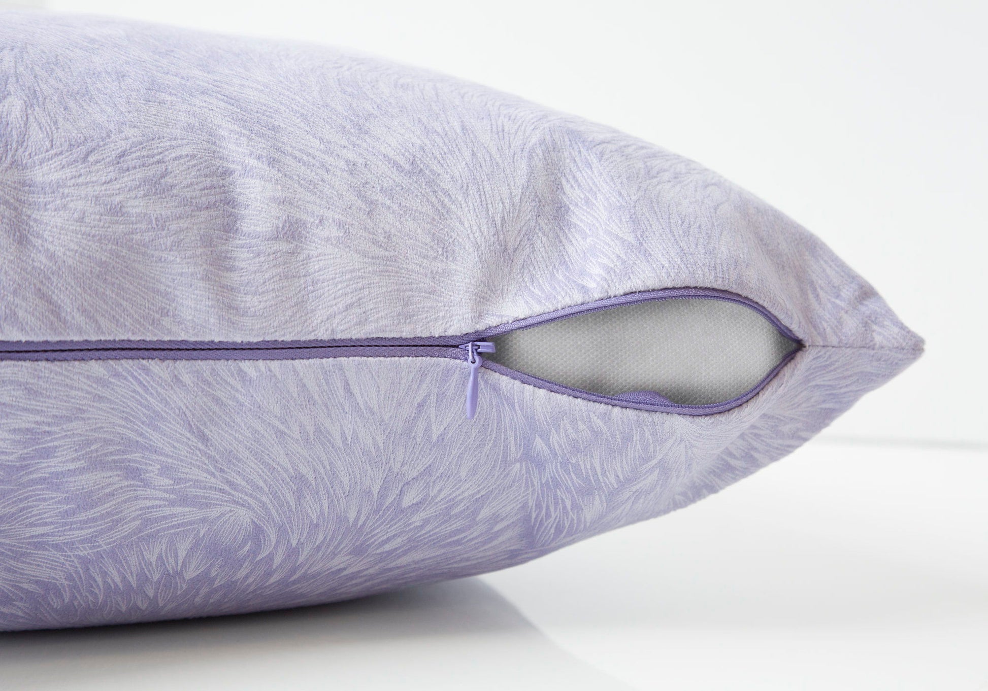 Close-up of a modern square decorative throw pillow in light lavender with a textured pattern, featuring a partially open purple zipper and revealing the hypoallergenic white insert included.