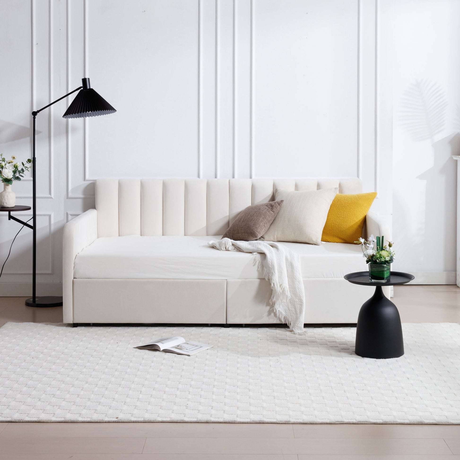 A modern living room featuring the Flora Upholstered Daybed with 2 drawers, ribbed tufted backrest in a lavish modern design, assorted throw pillows, a white blanket, a round black side table with flowers, black floor lamp, and textured white rug on light wood floors.