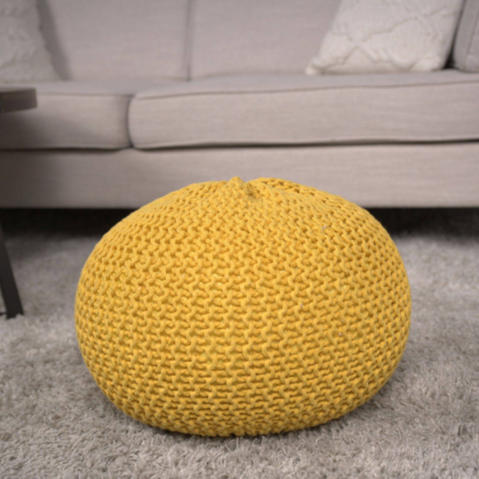 The Bordeaux Knitted Cotton Round Pouf in yellow rests on a light gray carpet before a beige sofa with textured cushions, adding cozy style to the living room as both ottoman and floor cushion.