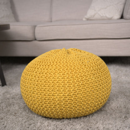 The Bordeaux Knitted Cotton Round Pouf in yellow rests on a light gray carpet before a beige sofa with textured cushions, adding cozy style to the living room as both ottoman and floor cushion.