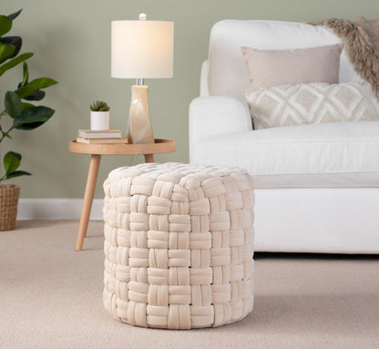 The Braided - Versatile Round Ottoman in cream sits on a beige carpet in the living room, near a white sofa with neutral pillows, a round wooden side table with a lamp, and a potted plant in the background.