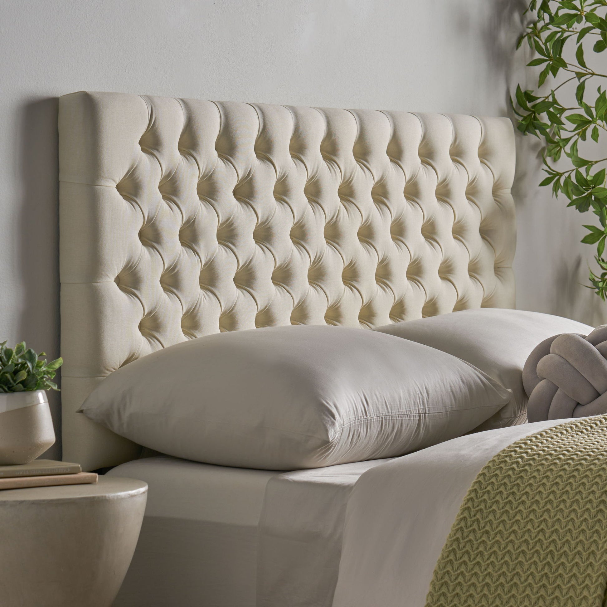 A close-up of the Annecy - Contemporary Upholstered Headboard in beige behind a neatly made bed with pale bedding, accented by potted plants and a green textured blanket for a serene bedroom setting.
