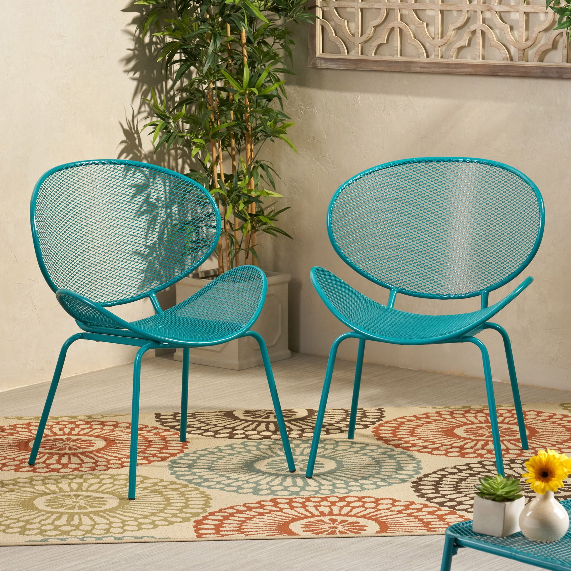 Two Elba - Contemporary Chair Set For Outdoor Relaxation in turquoise metal sit on a circular patterned rug, beside a small table with flowers and a potted plant, against a beige wall adorned with a decorative panel.