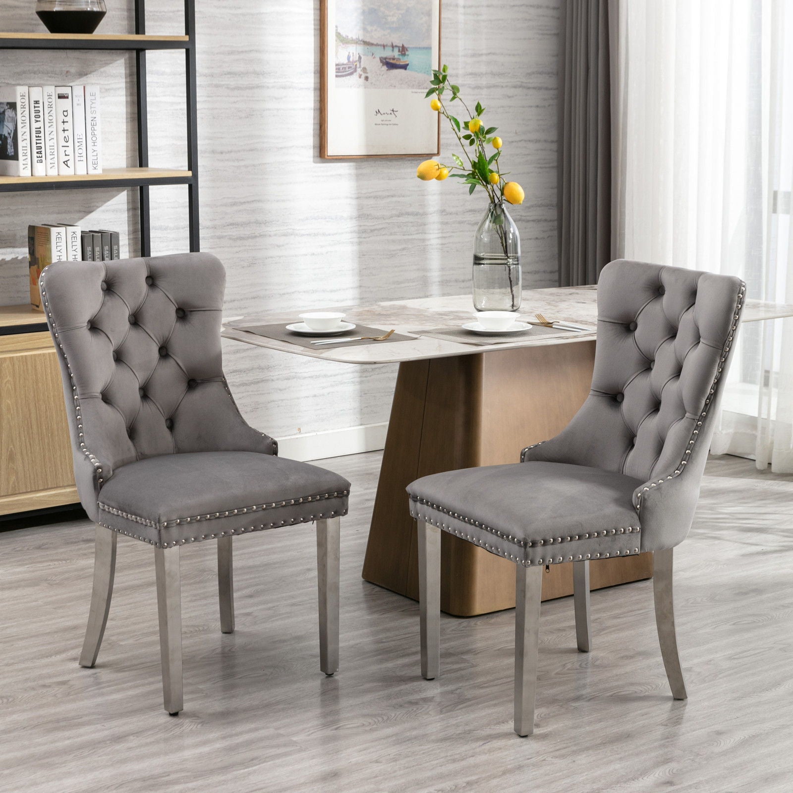 A set of two Nikki modern tufted velvet dining chairs with chrome legs and nailhead trim are arranged in a stylish dining room with wood floors, a marble top table, yellow flowers, bookshelves, and sheer-curtained windows.