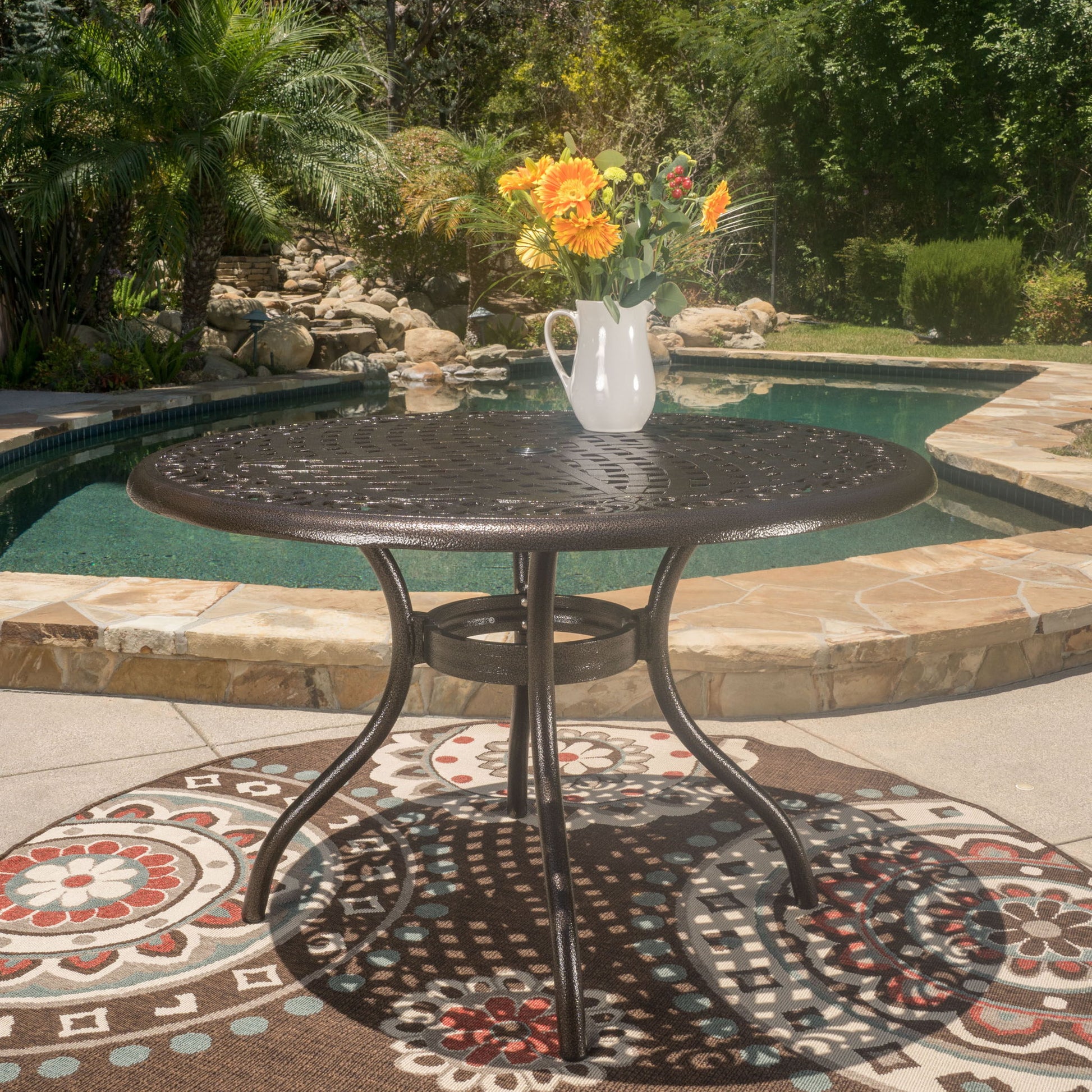 The Tranquilius Outdoor Round Dining Table with a lattice tabletop, umbrella hole, and curved legs sits on a patterned outdoor rug by the pool, featuring a white pitcher of yellow and orange flowers. Lush greenery and rocks surround the pool in the background.