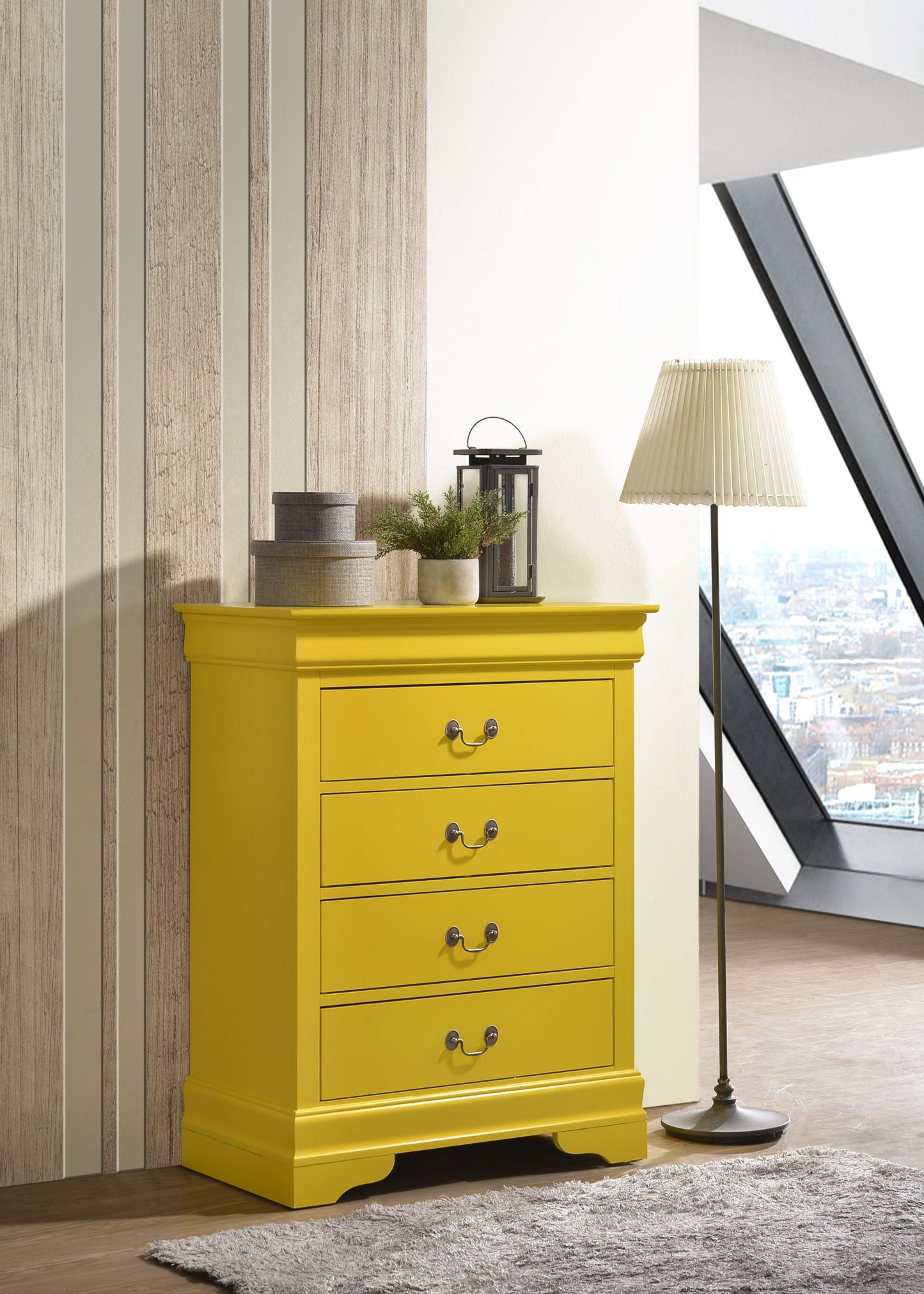 The Traditional Style Storage Chest Elegant in bright yellow, featuring four drawers, stands on a light rug next to a floor lamp with a pleated shade. Decorative accents and a lantern top the chest, with city views visible through large windows behind.