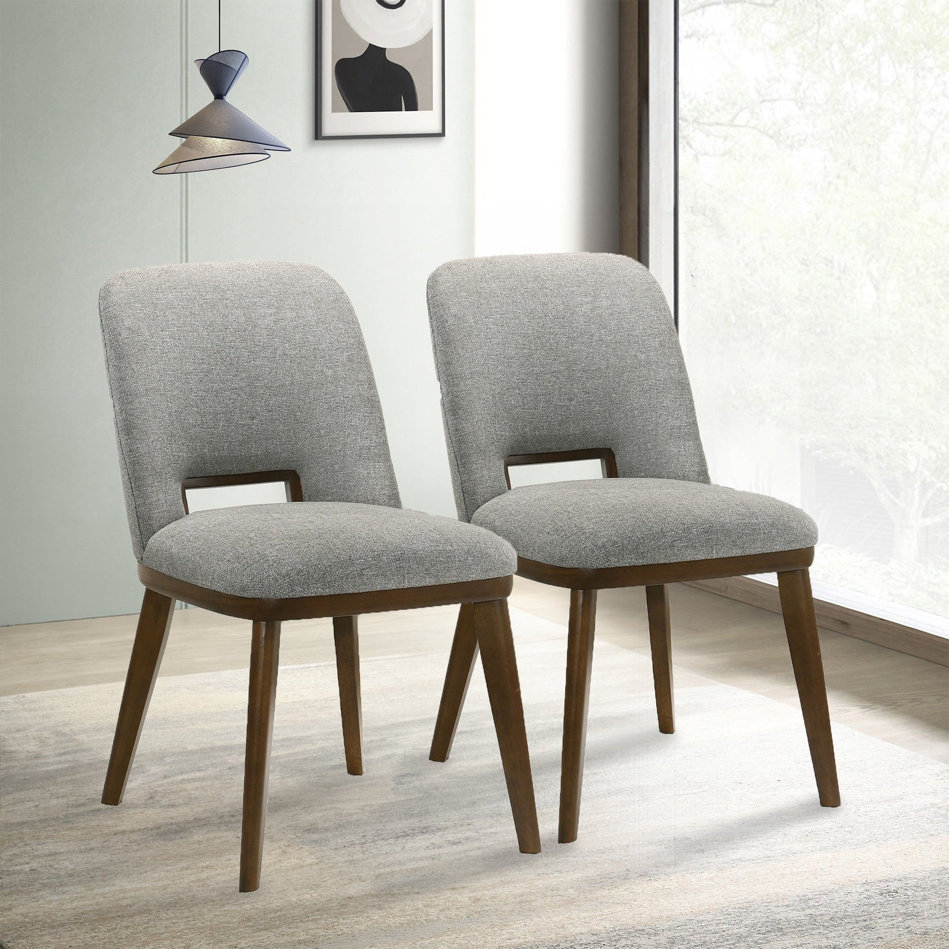 Two Blake - Modern Dining Chairs (Set of 2) with gray upholstery and wooden legs are placed side by side on a light rug in a modern room featuring large windows, minimalist decor, and a pendant light overhead.
