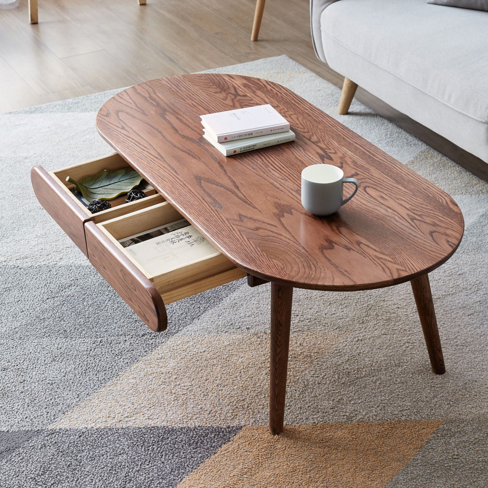 The Capsule Centre Low Table With Drawers Coffee Table, featuring angled legs and an open side drawer with small items, holds a white mug and two stacked books. It stands on a patterned rug beside a gray sofa.