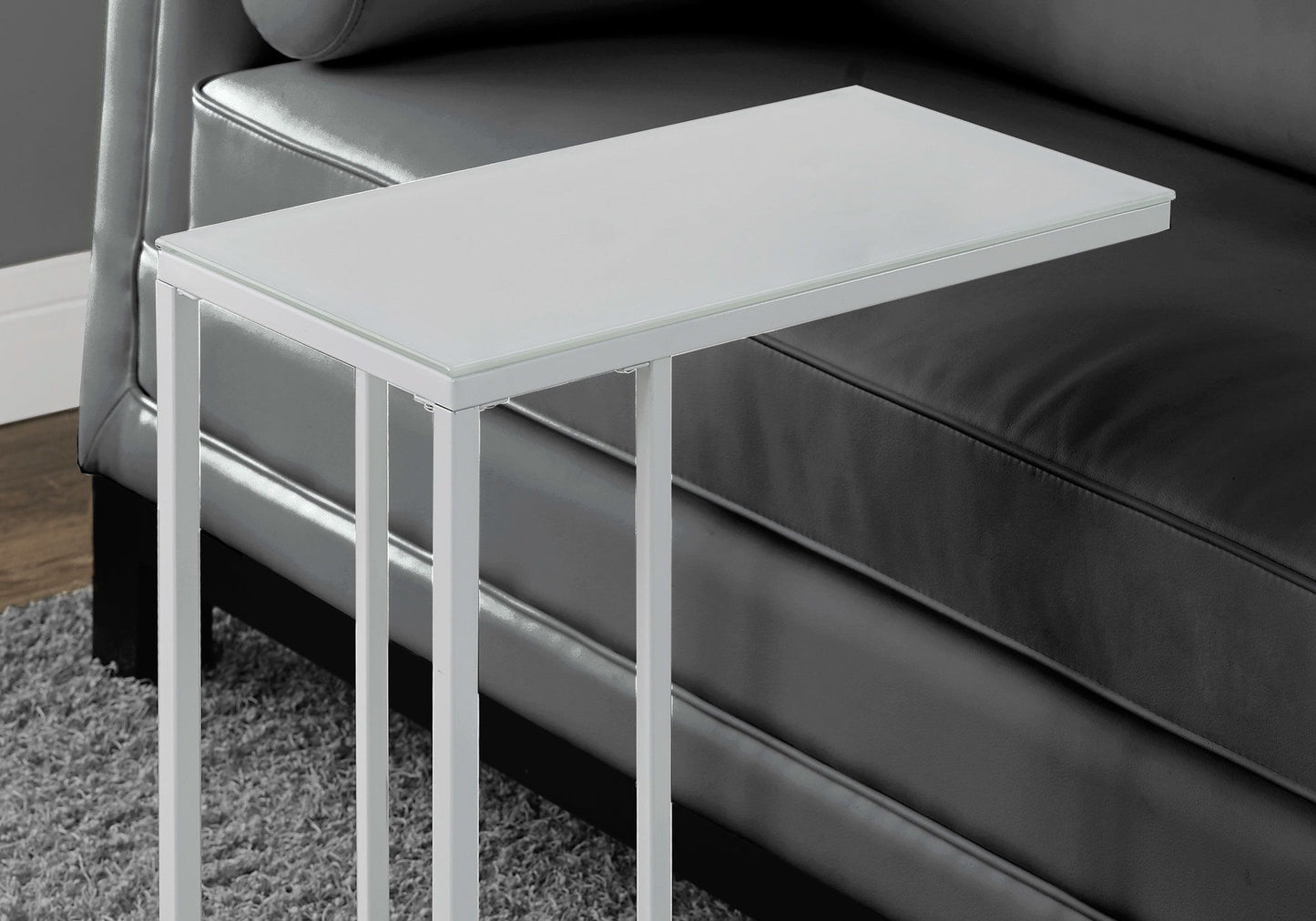 A C-shaped Accent Table with a tempered glass top and stylish contemporary design stands beside a dark gray leather sofa on a gray carpet, its minimalist metal frame positioned near the sofa armrest.